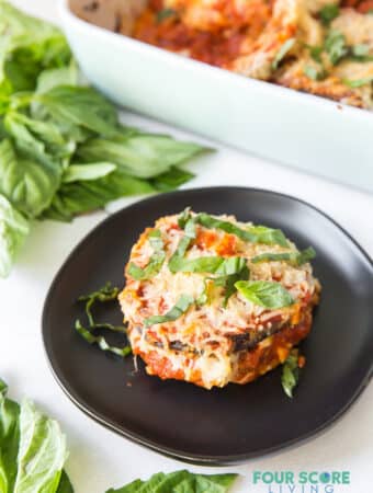 A serving of eggplant parmesan on a black plate next to a casserole pan.