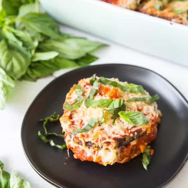 A serving of eggplant parmesan on a black plate next to a casserole pan.