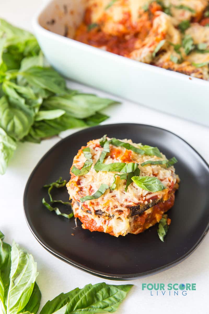 A serving of eggplant parmesan on a black plate next to a casserole pan.