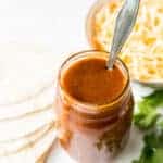 a jar of enchilada sauce with a spoon it in, next to a bowl of cheese, a stack of tortillas and some cilantro.