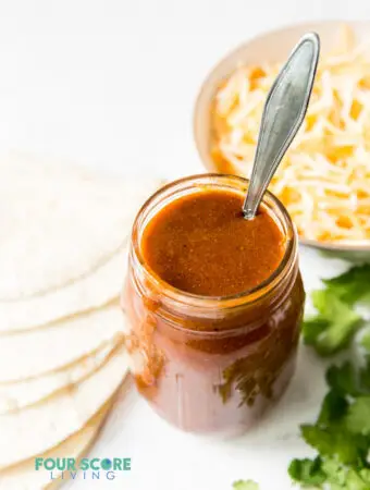 a jar of enchilada sauce with a spoon it in, next to a bowl of cheese, a stack of tortillas and some cilantro.