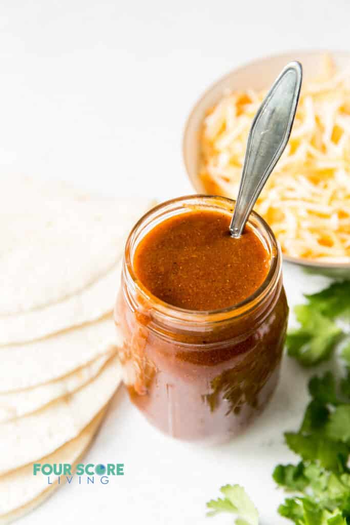 a jar of enchilada sauce with a spoon it in, next to a bowl of cheese, a stack of tortillas and some cilantro.