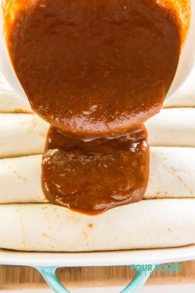 Enchilada sauce being poured over a pan of rolled enchiladas.