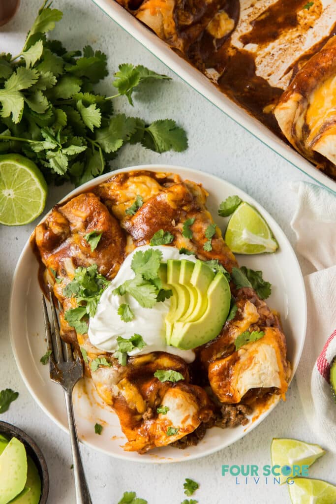 two enchiladas on a plate topped with sour cream, avocado, and cilantro.