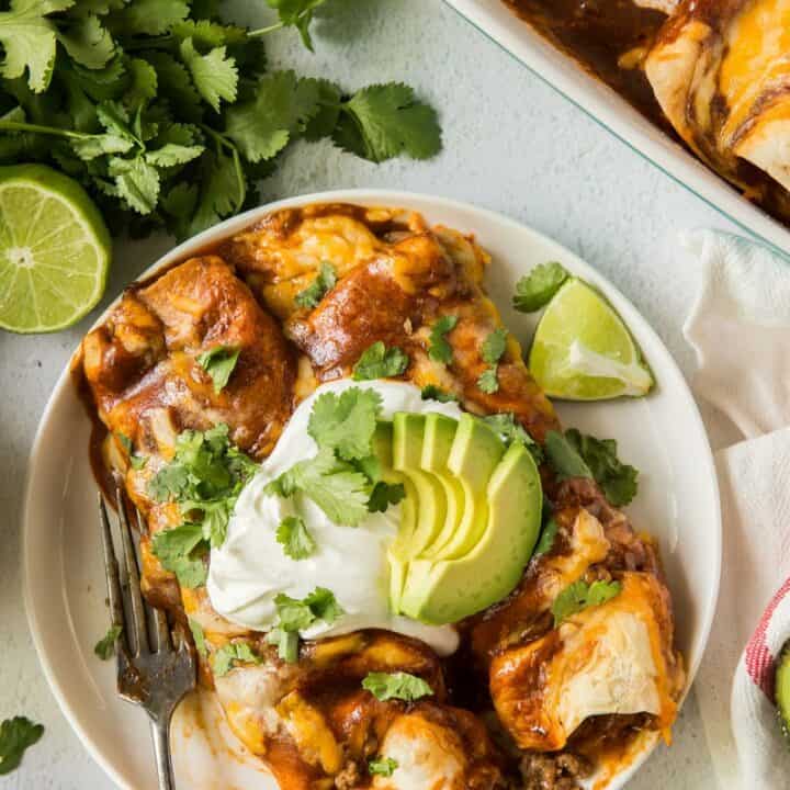 two enchiladas on a plate topped with sour cream, avocado, and cilantro.