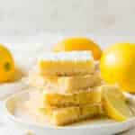 a plate of 4 lemon bars, stacked on top of each other, surrounded by lemons.