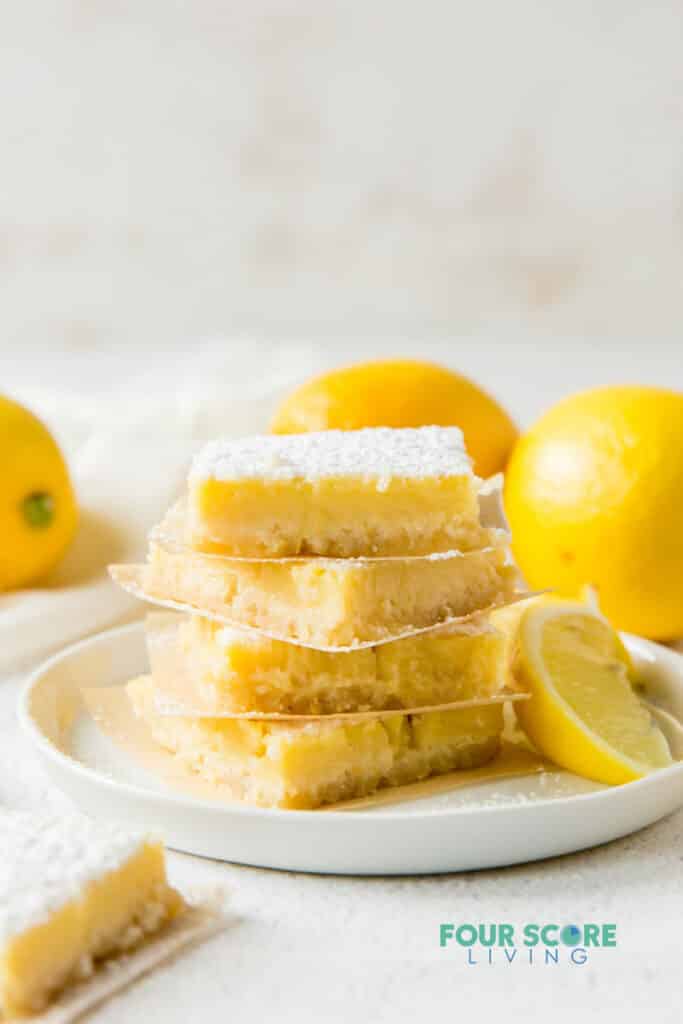 a plate of 4 lemon bars, stacked on top of each other, surrounded by lemons.