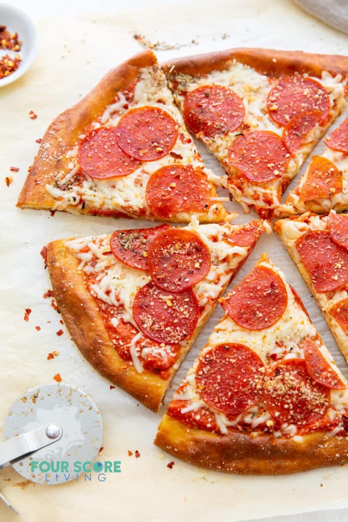 Pepperoni and cheese pizza sliced into wedges on parchment paper with a pizza slicer and a small white dish of red pepper flakes in the background