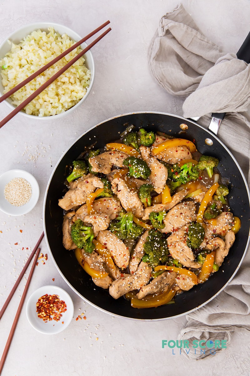 top down view of a chicken and broccoli stir fry in a frying pan, surrounded by chopsticks, rice, and garshishes.