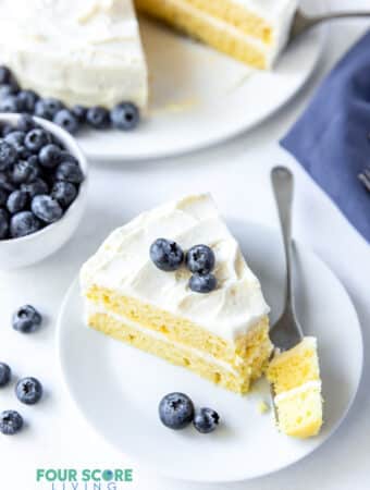 a slice of vanilla layer cake on a plate with a bit being taken. Cake is topped with blueberries.