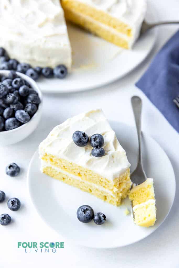 a slice of vanilla layer cake on a plate with a bit being taken. Cake is topped with blueberries.