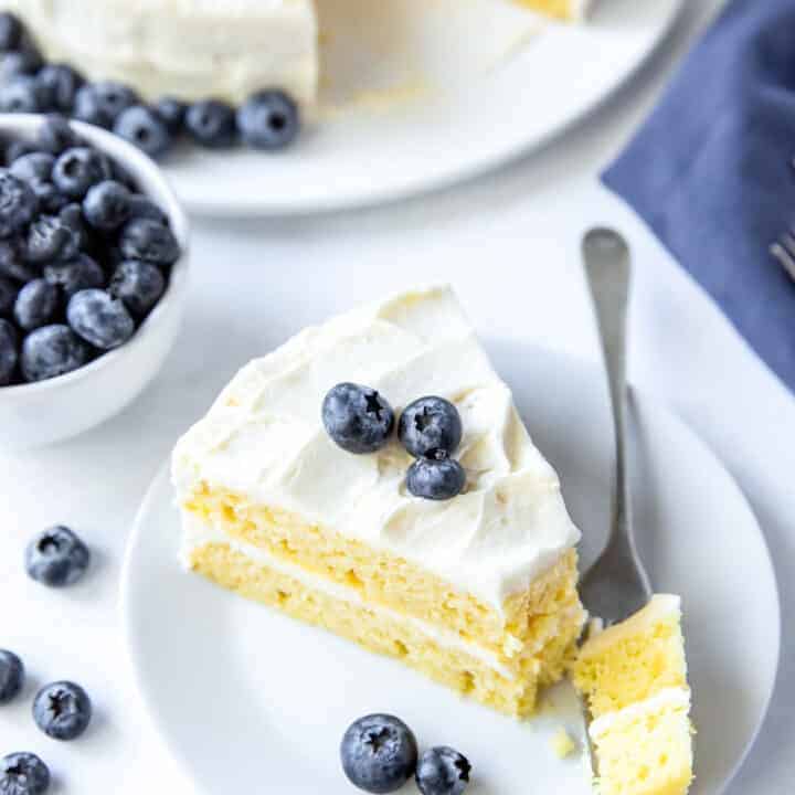 a slice of vanilla layer cake on a plate with a bit being taken. Cake is topped with blueberries.