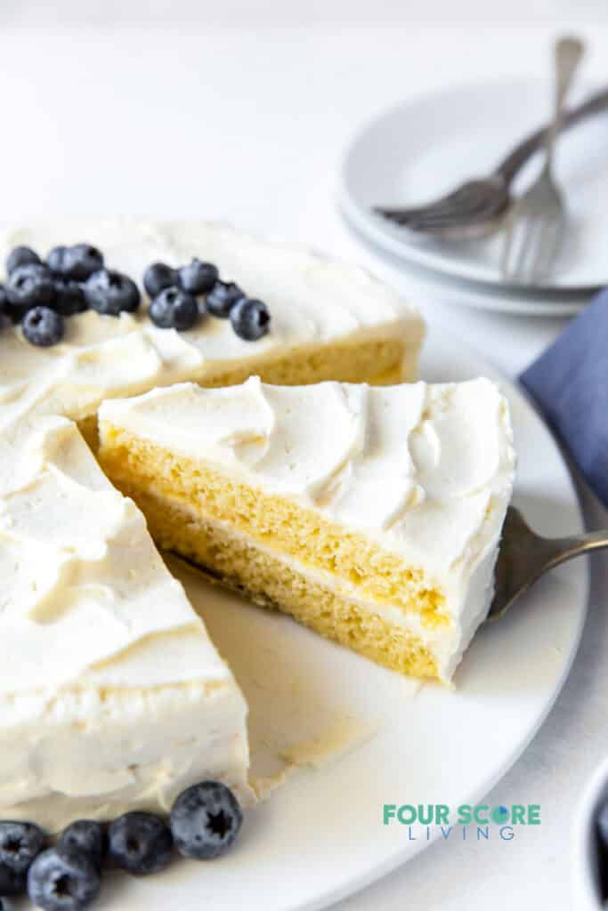 a round two layer cake with vanilla frosting topped with blueberries. A slice of cake is being served.