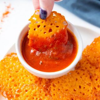 a plate of keto cheese chips with a hand dipping one chip into a cup of marinara sauce