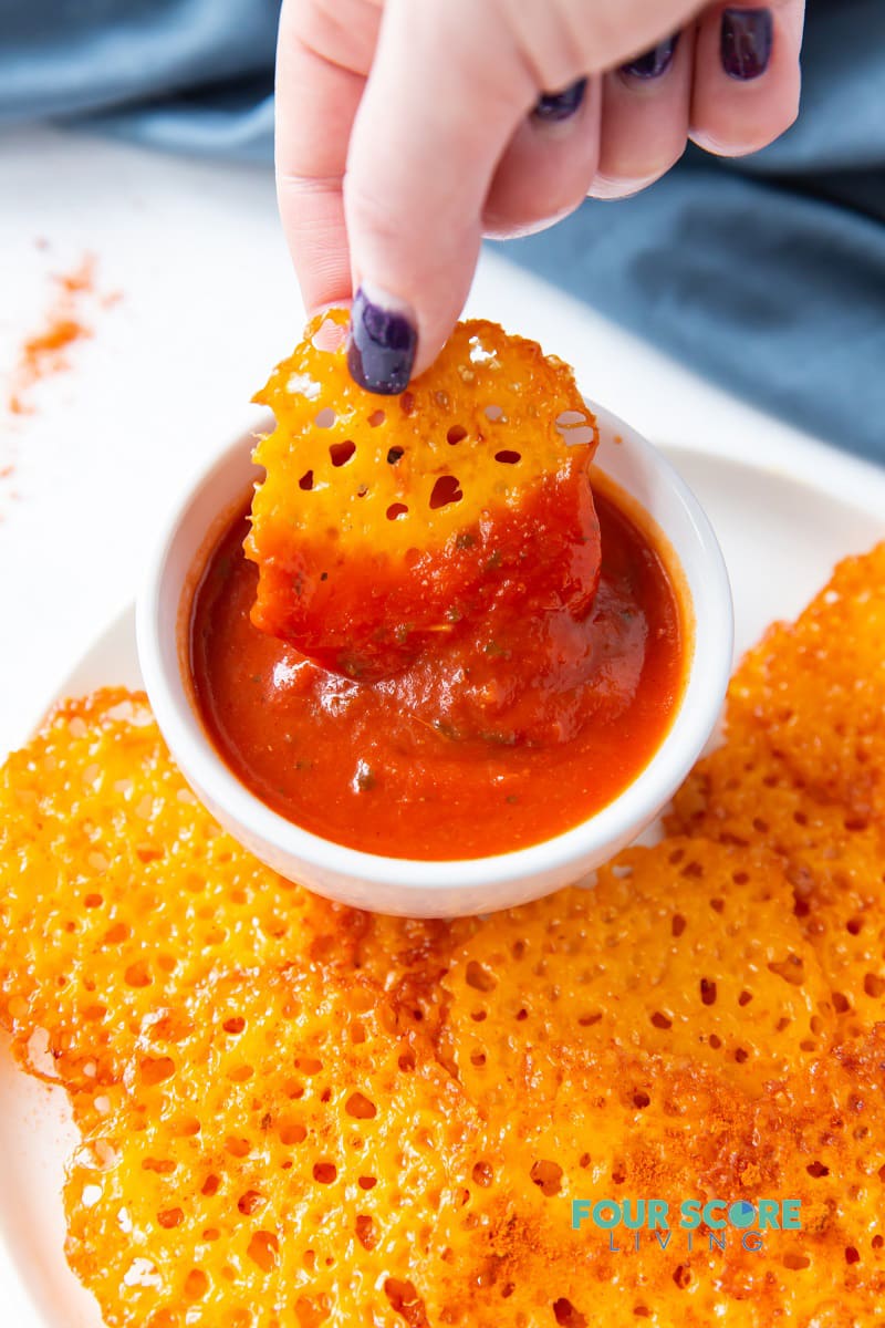 a plate of keto cheese chips with a hand dipping one chip into a cup of marinara sauce