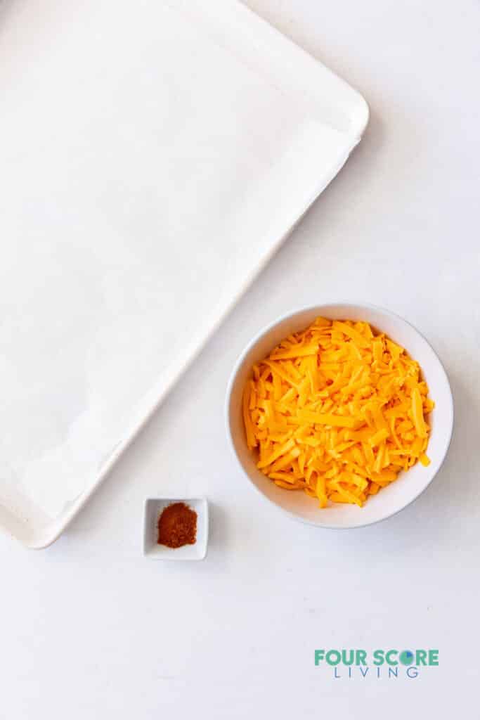 Ingredients needed to make keto cheese chips, including cheese and seasonings, next to a parchment lined square plate a