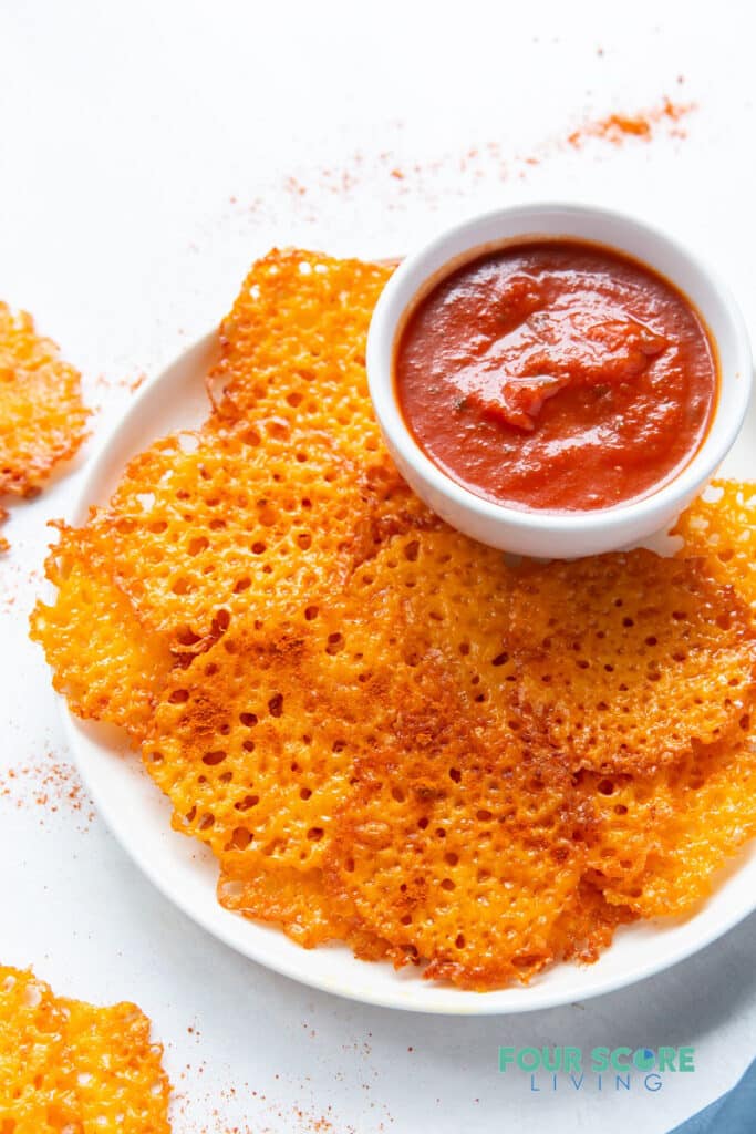 a round white plate of baked cheese chips with a cup of marinara sauce