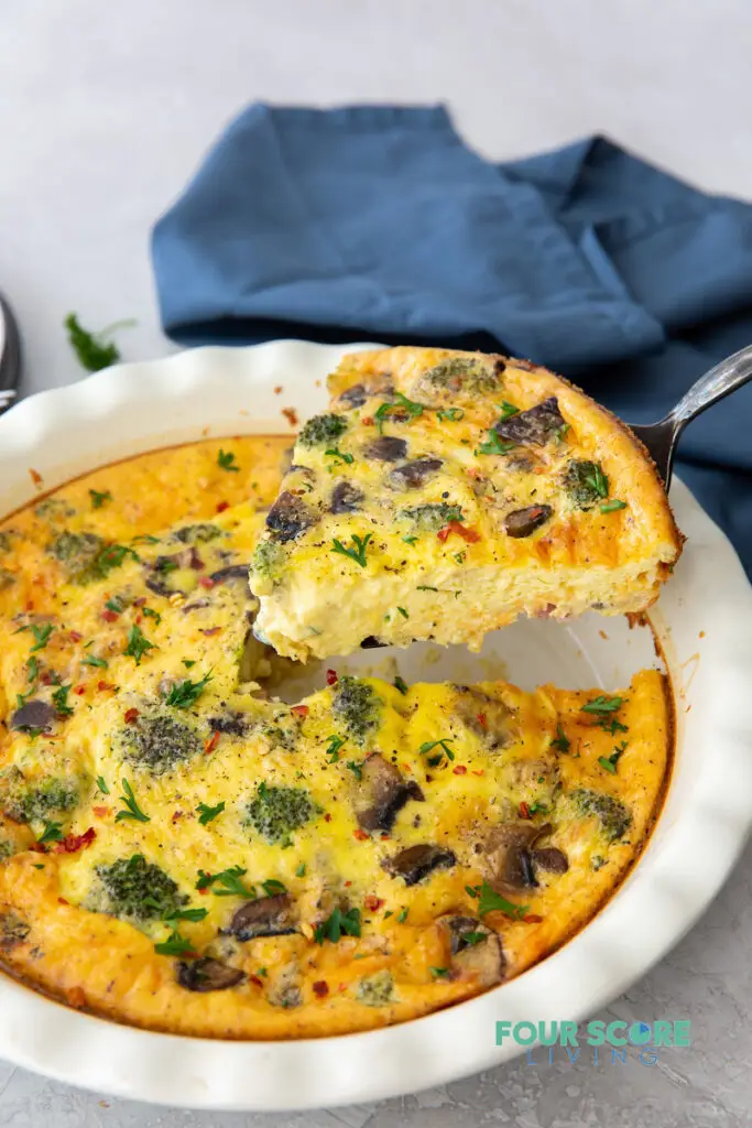 a white ceramic pie pan of quiche with broccoli and mushrooms, one wedge is being lifted out.