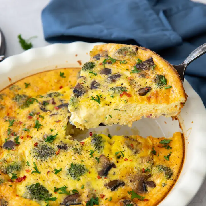 a white ceramic pie pan of quiche with broccoli and mushrooms, one wedge is being lifted out.