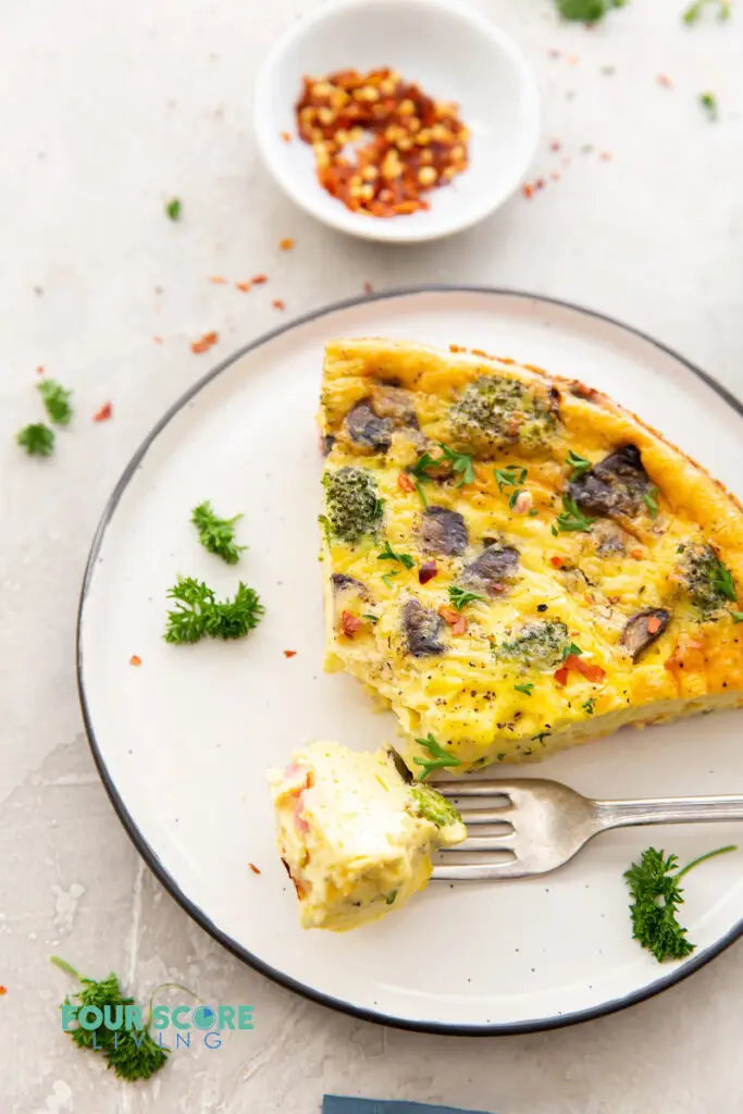 a white plate with a black rim with a large slice of quiche on it. Garnished with red pepper flakes and parsley.