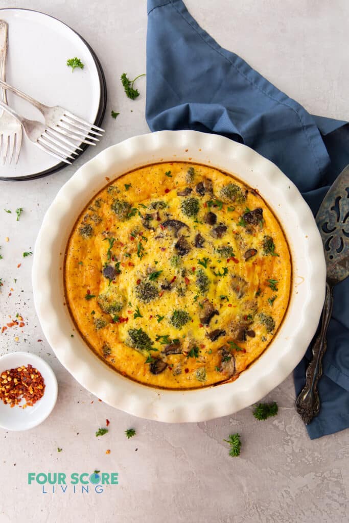 top down view of a quiche in a white ceramic pie pan next to a white plate with three forks, a pie server, and a blue napkin.