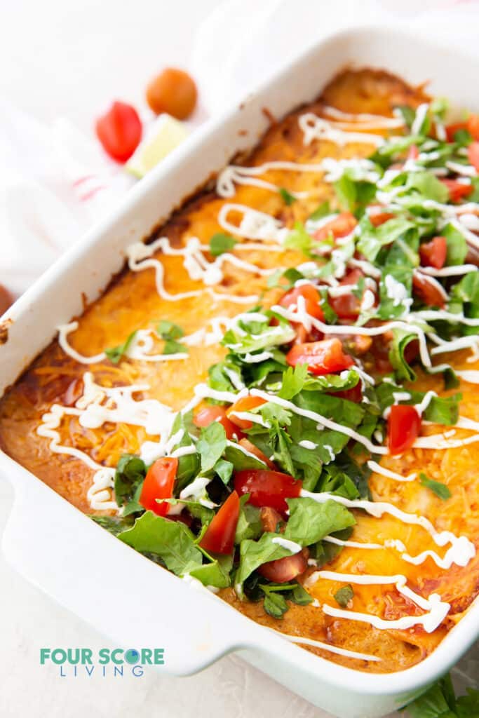 a white rectangular casserole dish on an angle, filled with taco casserole and topped with drizzled sour cream, lettuce, and tomatoes