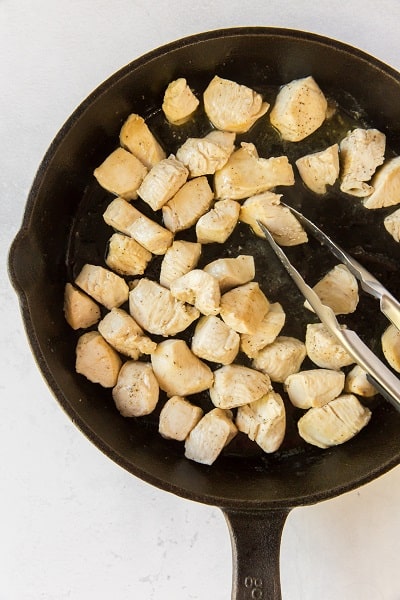 Top view photo of a cast iron skillet with cubed chicken frying until browned on the edges.