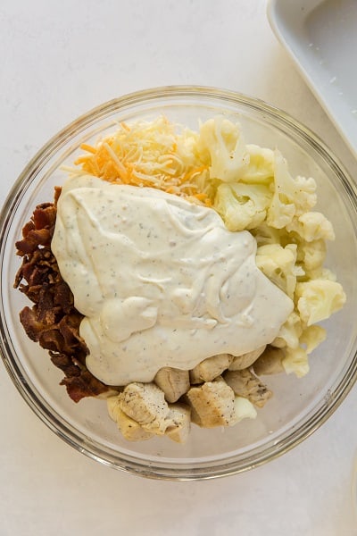Top view photo of a large glass bowl with bacon, ranch mixture, cheese, chicken, and cauliflower, ready to be mixed together.