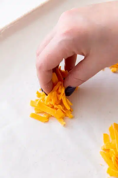 Photo of a hand placing shredded cheddar cheese into a little stack on a parchment lined baking sheet.