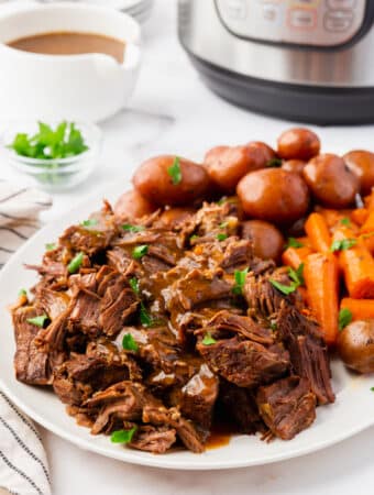 a plate of pot roast and gravy, baby potatoes and carrots in front of an instant pot