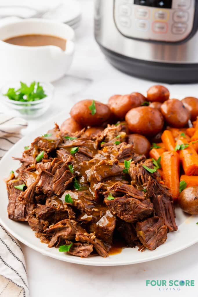 a plate of pot roast and gravy, baby potatoes and carrots in front of an instant pot