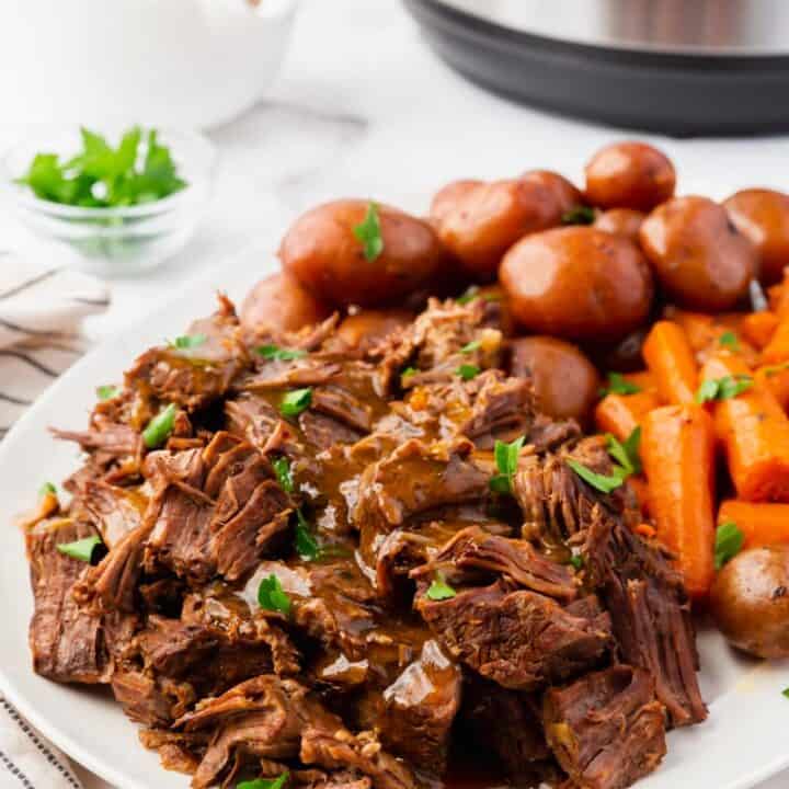 a plate of pot roast and gravy, baby potatoes and carrots in front of an instant pot