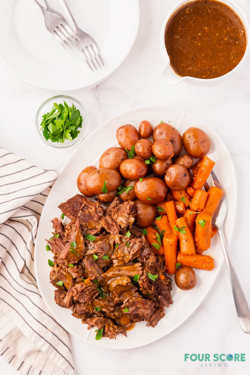 a platter of pot roast, carrots, and baby potatoes. To the side is a large gravy boat filled with beef gravy.