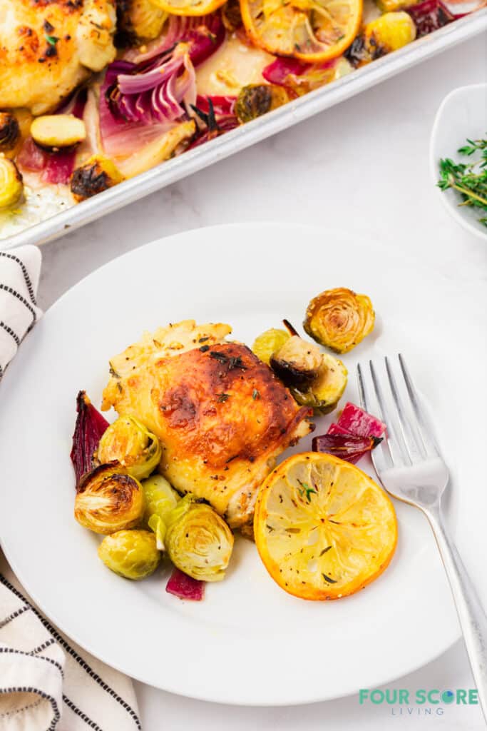 a white plate with a roasted chicken thigh, brussels sprouts, and a roasted lemon.