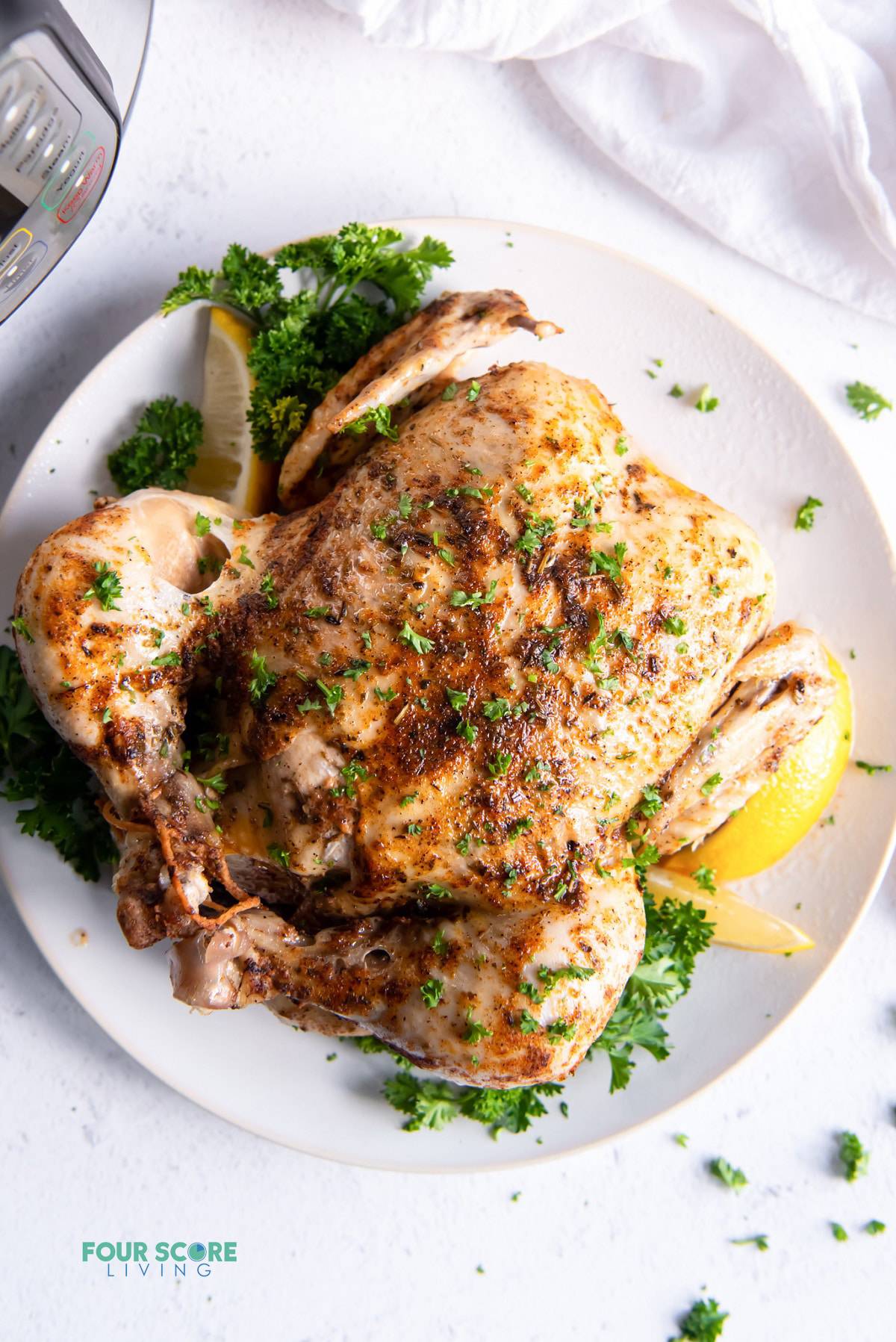 a whole chicken seasoned with herbs and lemon on a round white platter next to an instant pot.