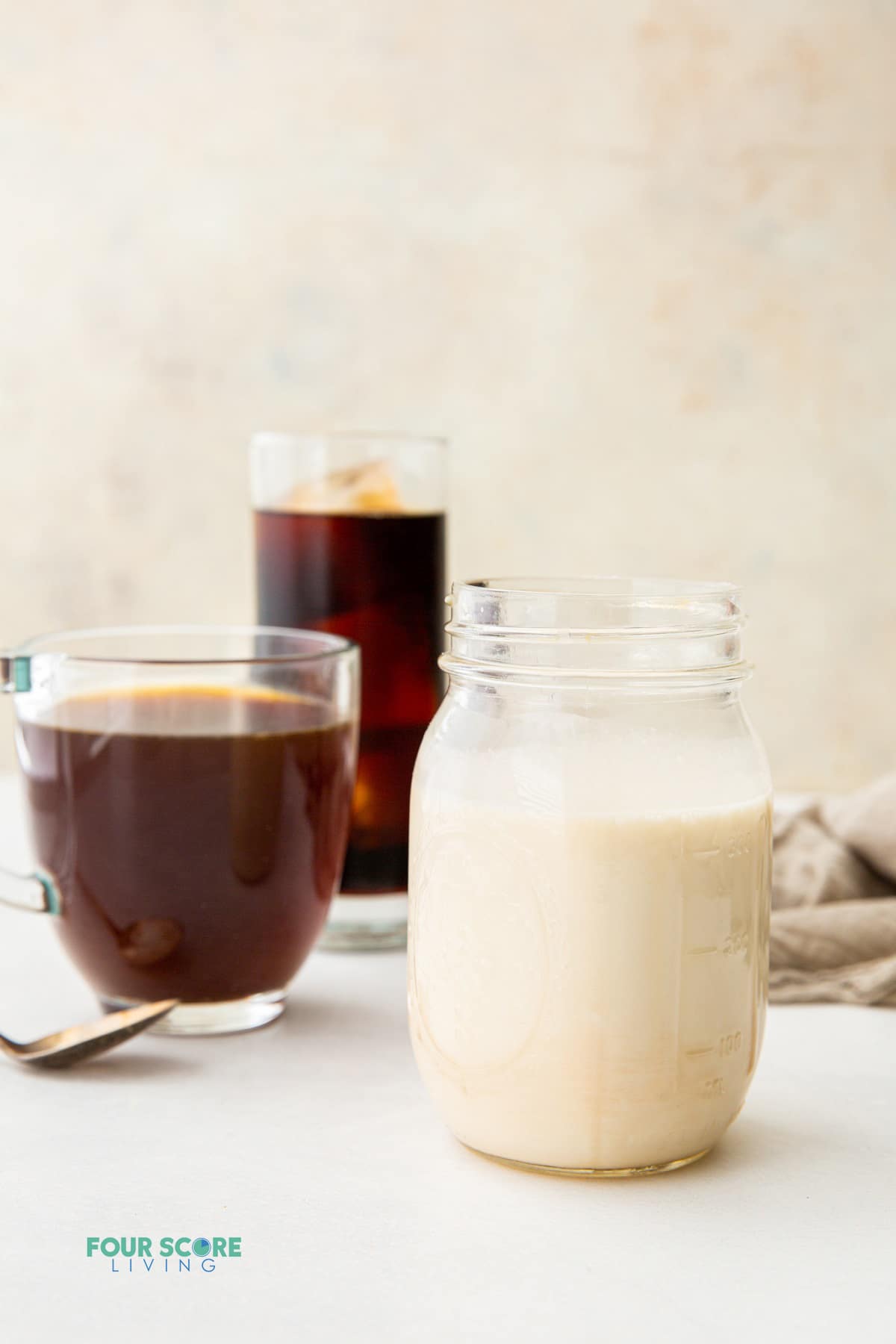 a mason jar filled with homemade almond milk creamer in front of a glass mug of coffee and a tall glass of iced coffee.