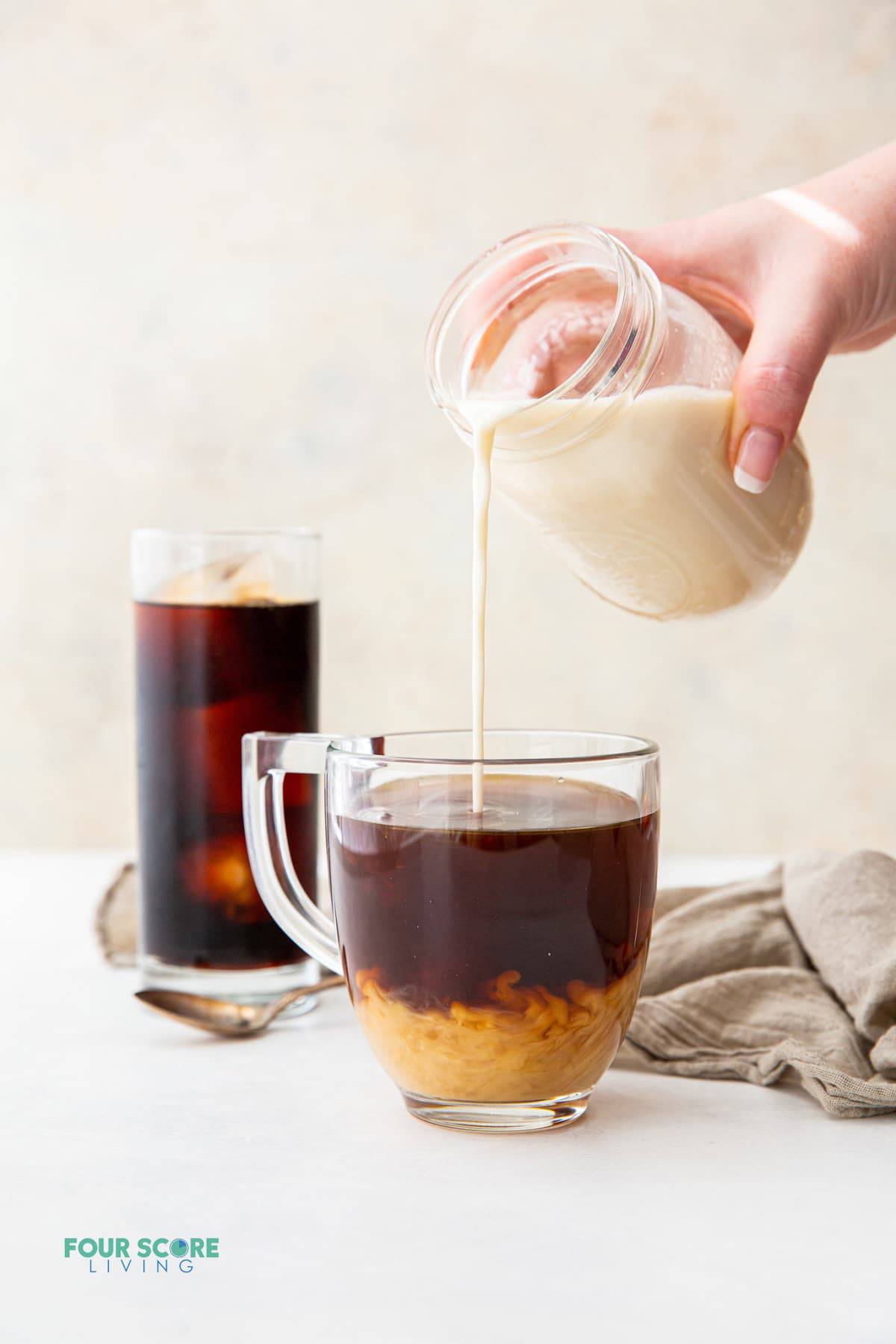 a glass mug of hot coffee with homemade keto creamer being poured in from a mason jar. A clear glass of black iced coffee is in the background