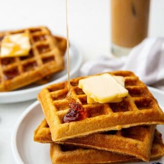 a plate of three square keto waffles topped with two pats of butter. Syrup is being poured on top. an iced coffee is in the background.