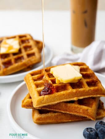 a plate of three square keto waffles topped with two pats of butter. Syrup is being poured on top. an iced coffee is in the background.