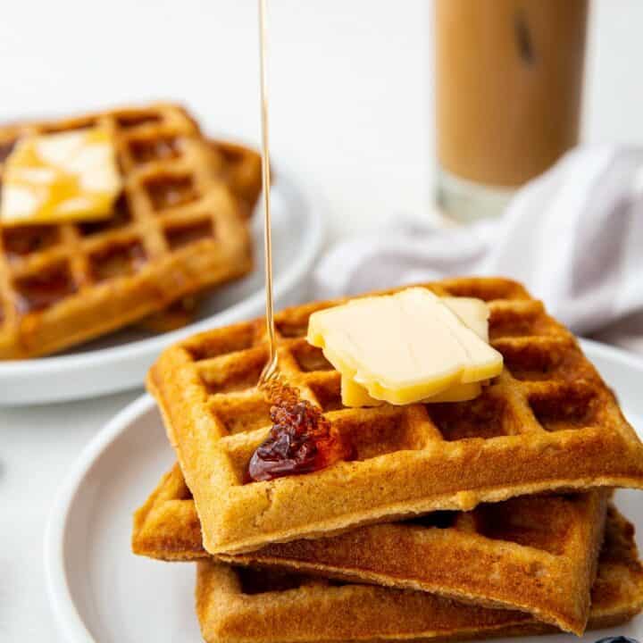 a plate of three square keto waffles topped with two pats of butter. Syrup is being poured on top. an iced coffee is in the background.