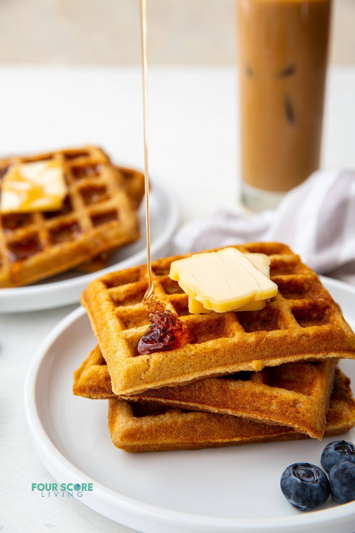 a plate of three square keto waffles topped with two pats of butter. Syrup is being poured on top. an iced coffee is in the background.