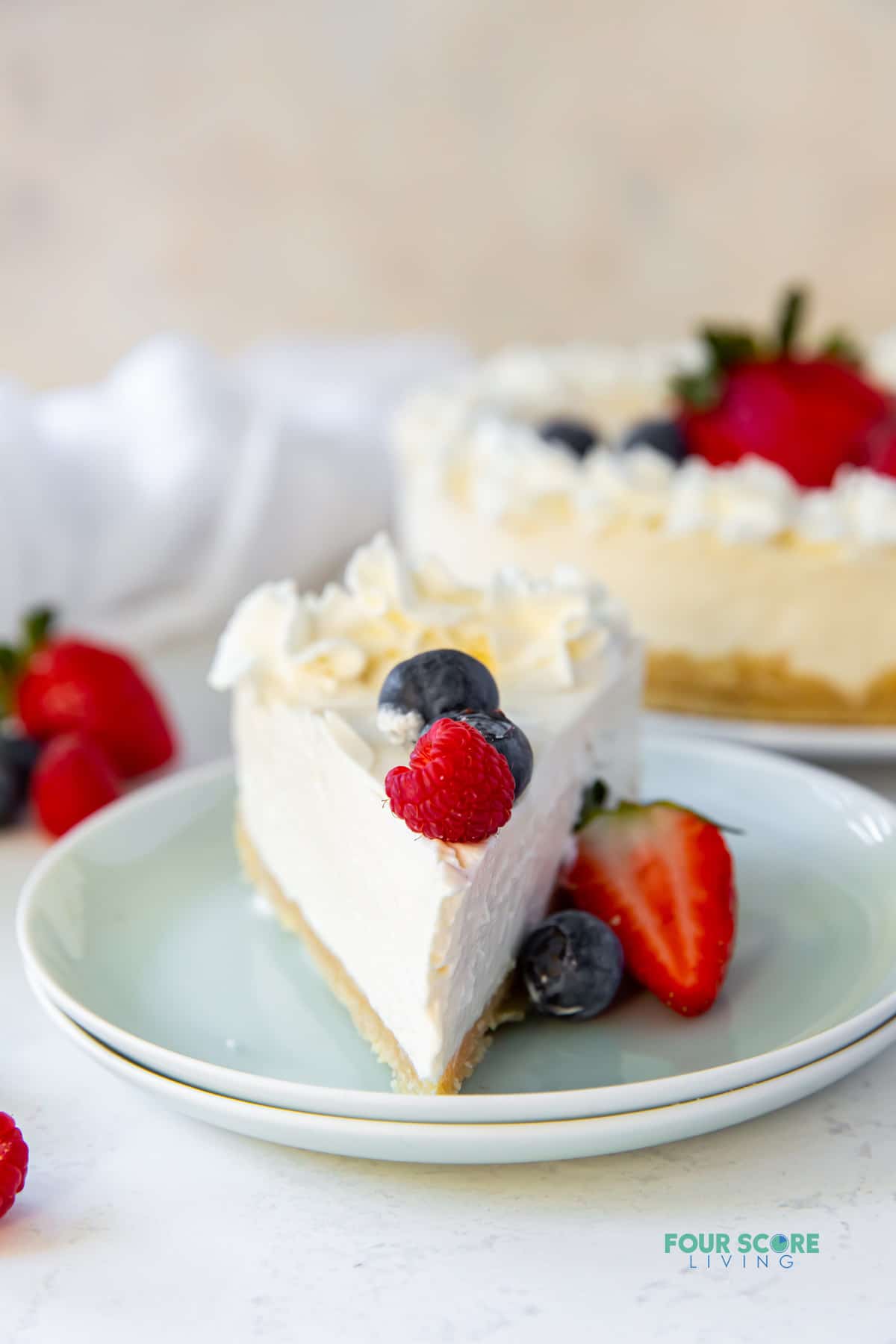 a slice of no-bake cheesecake with the point facing you on a pale green doubled plate. The slice is topped with fresh berries and whipped cream