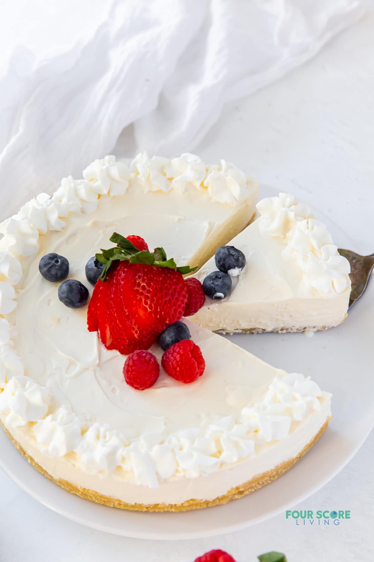 a whole keto no bake cheesecake with whipped cream topping and fresh berries in the center. A piece is gone, and the next piece is being removed with a cake server