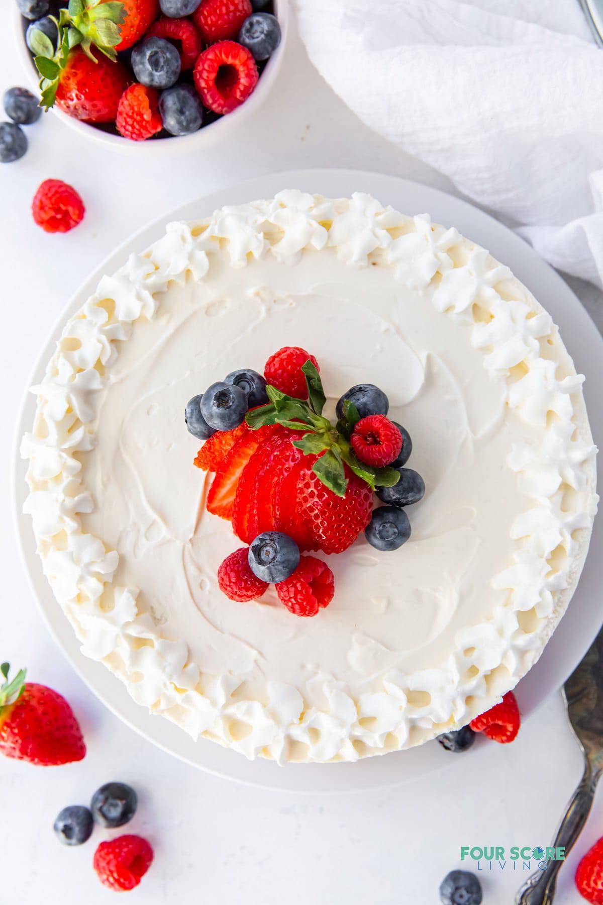 a whole keto no bake cheesecake. the top is decorated with whipped cream around the edges and fresh berries piled in the center. Image is taken from above