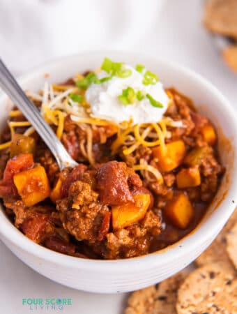 a white bowl filled with chipotle chilli made with beef, sweet potato, and tomatoes and topped with cheese and sour cream