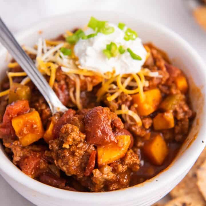 a white bowl filled with chipotle chilli made with beef, sweet potato, and tomatoes and topped with cheese and sour cream