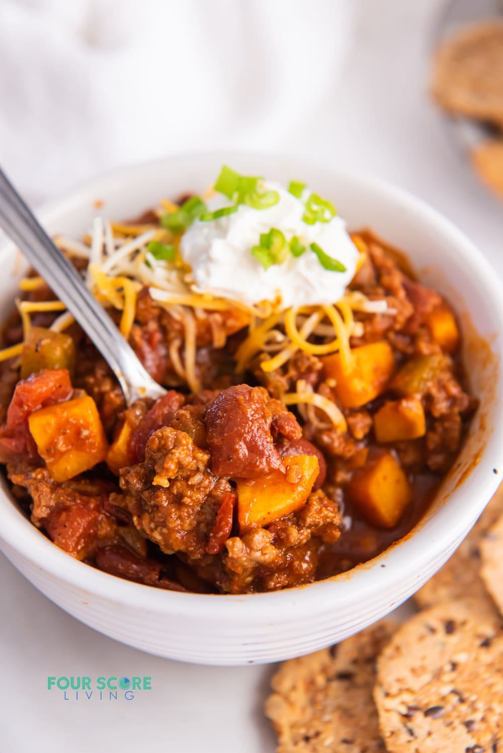 a white bowl filled with chipotle chilli made with beef, sweet potato, and tomatoes and topped with cheese and sour cream