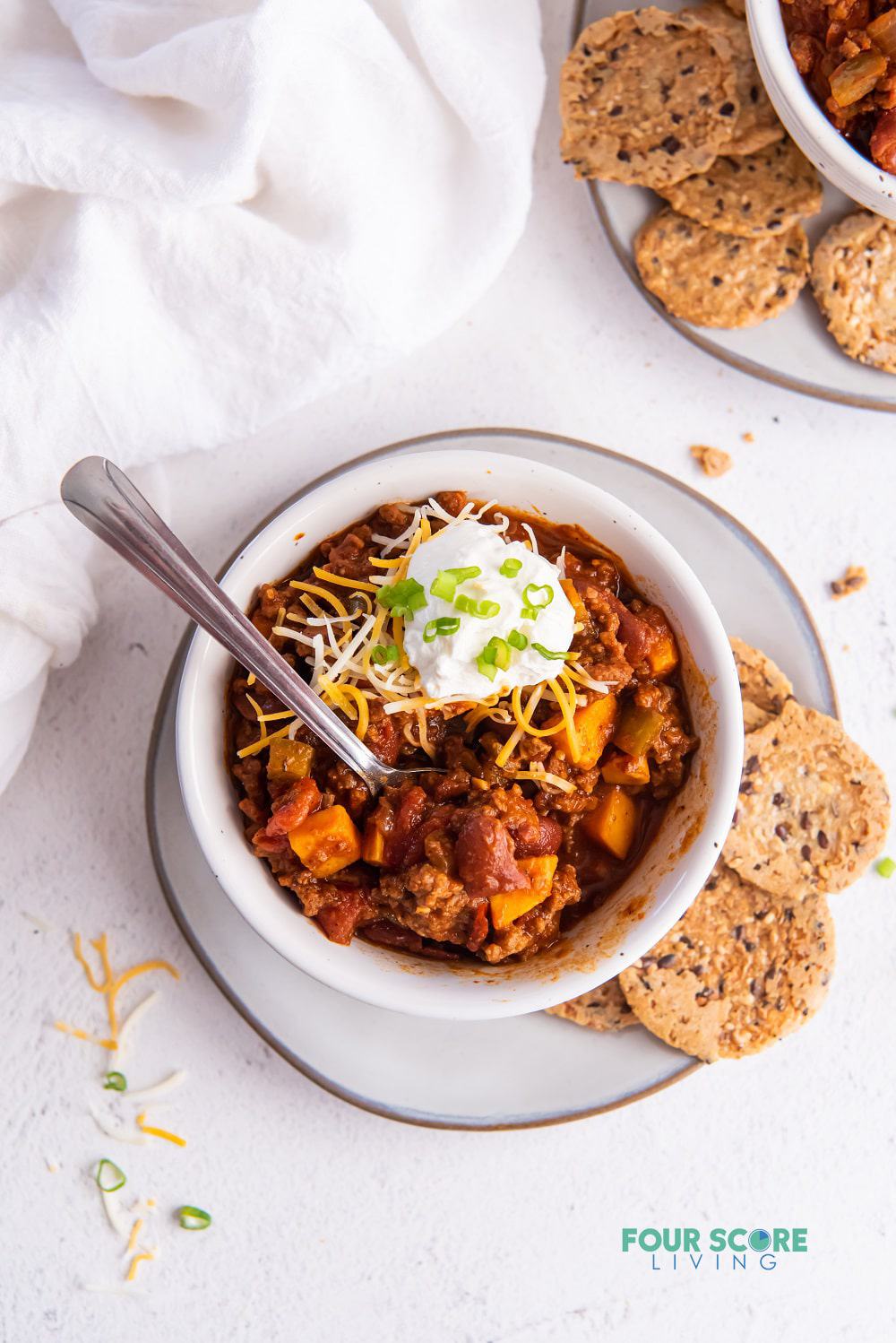 a bowl of chili topped with cheese and sour cream on a plate with seed crackers on the side.