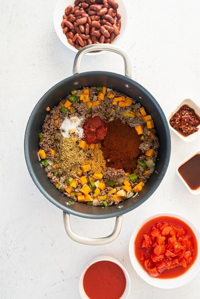 Top view photo of a large stock pot with tomato paste, chili powder, cumin, oregano, salt, and pepper added to the chili base.