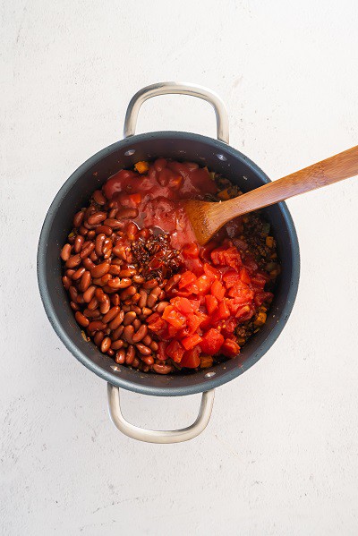Top view photo of a large stockpot with tomatoes, tomato sauce, kidney beans, chipotle peppers, and chipotle sauce added to the chili, and stirred together with a wooden spoon.